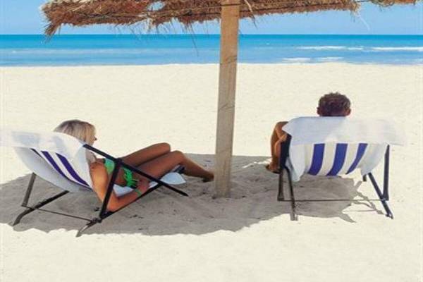 Zwei Personen sitzen unter einem Strohhut auf dem Strand, einer liest ein Buch.