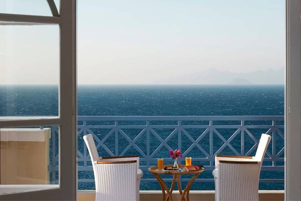 Aussicht vom Balkon des Mitsis Summer Palace mit zwei weißen Stühlen, einem Tisch mit Getränken und einer Aussicht auf das Meer und die Berge.