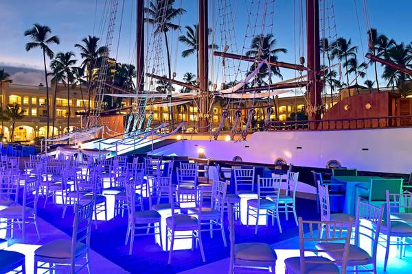 Abendliche Feier auf der Terrasse des JOIA Bávaro by Iberostar Hotel mit blau beleuchteten Stühlen und einem Schiff im Hintergrund.
