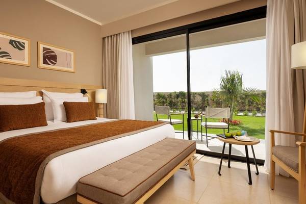 Hotelzimmer des Jaz Elite Amara mit einem großen Bett, zwei Nachttischen, einer Bank, einem kleinen Tisch mit Obst und einem Balkon.