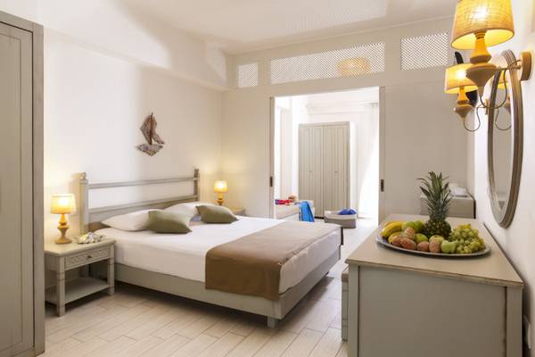 Innenansicht eines Zimmers im Almyrida Beach Hotel mit einem Bett, Nachttischen und Früchten auf einem Tisch.