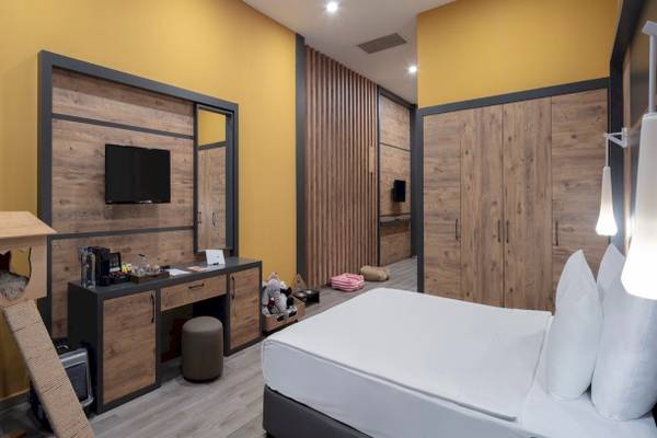 Hotelzimmer im Nirvana Cosmopolitan mit gelben Wänden, Holzelementen, einem Fernseher, Schreibtisch, Bett und Katzenzubehör.