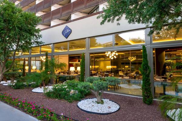 Außenansicht des Nirvana Cosmopolitan Hotels mit Garten, Bäumen und Glasfassade.