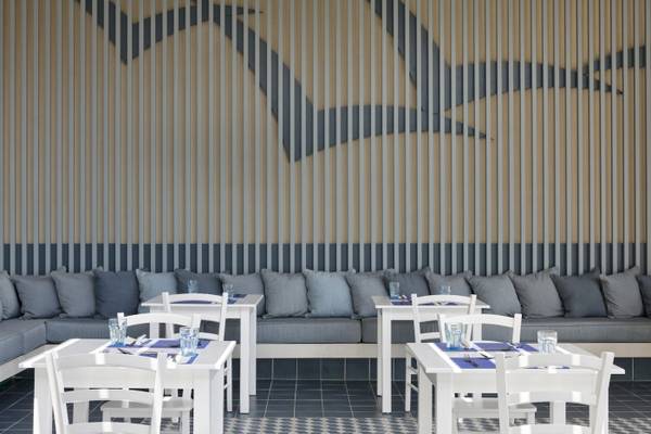 Restaurantbereich im Atlantica Kalliston Resort mit weißen Tischen und Stühlen, grauen Kissen und blauen Wanddekorationen.