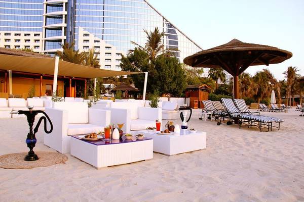 Loungebereich am Strand des Jumeirah Beach Hotels mit weißen Möbeln und einem großen Gebäude im Hintergrund.