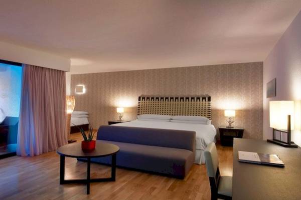 Innenansicht eines Hotelzimmers im Sheraton Rhodes Resort mit Bett, Schreibtisch, Sesseln und Pflanzen.