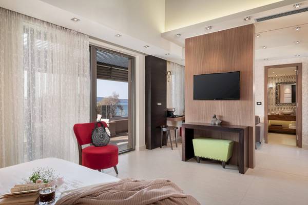 Innenansicht eines Zimmers im Cretan Dream Resort & Spa mit Bett, rotem Stuhl, Fernseher und Aussicht auf das Meer.