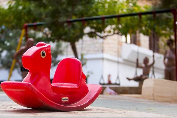 Ein rotes Schaukelpferd im Freien, im Hintergrund sind Kinder auf Schaukeln zu sehen.