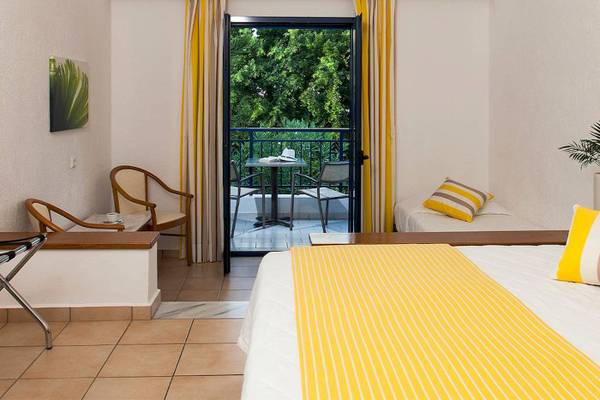 Hotelzimmer von Chrissi Amoudia mit Balkon, Bett, Tisch, Stühlen und Blick auf Bäume.