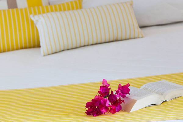 Foto von einem Bett im Chrissi Amoudia Hotel mit gelben Kissen und einer offenen Buch mit Blumen darauf.