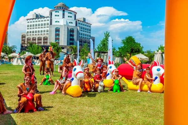 Gruppe von Kindern und Erwachsenen, die auf aufblasbaren Pferden und anderen Spielgeräten im Freien vor dem Melas Lara Hotel spielen.