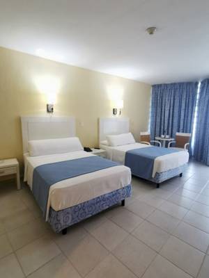 Zwei Doppelbetten im Zimmer des Muthu Playa Varadero Hotels, jedes mit weißen und blauen Laken, Nachttischen und Lampen an der Wand.