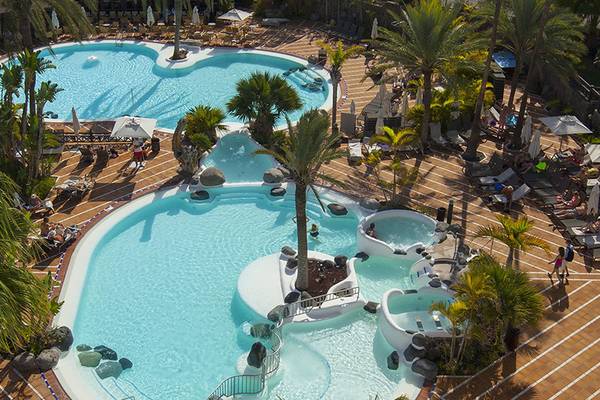 Oberflächenansicht des Schwimmbads im Corallium Beach Hotel mit mehreren Becken, Palmen und Sonnenliegen.