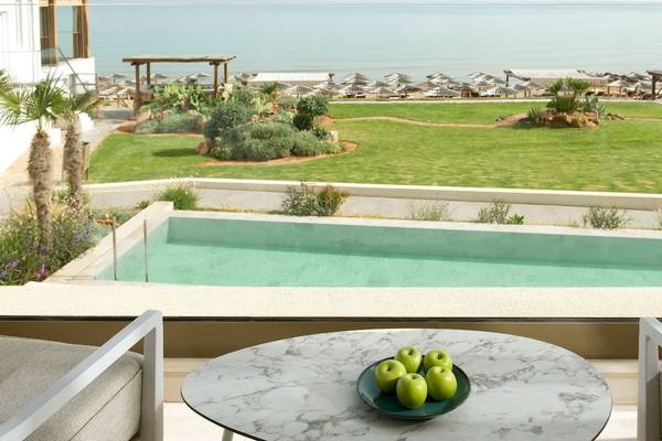 Blick vom Balkon des Mitsis Selection Rinela Hotels auf eine Terrasse mit Tisch, Äpfeln und Blick auf den Pool und das Meer.