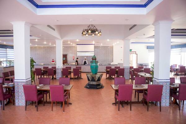 Innenbereich des Stella Gardens Resort & Spa Makadi Bay mit geometrischen Wandverkleidungen, runden Tischen, roten Stühlen, grünem Brunnen und hängender Lampe.