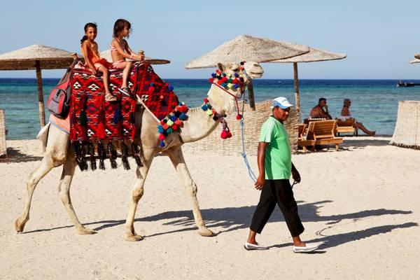 Zwei Kinder reiten auf einem Kamel am Strand, ein Mann führt das Kamel.