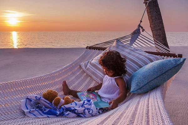 Ein kleines Maedchen liegt auf einem Hammock am Strand und liest ein Buch.