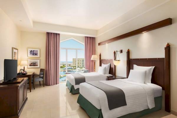 Doppelzimmer mit Meerblick im Wyndham Garden Salalah Mirbat Hotel, mit zwei Betten, Fernseher und Tisch, sowie ein großer Fenster mit Poolsicht.