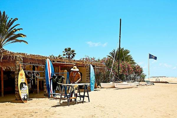 Ein Strand mit einem Surfshop, einem Mann, der an einer Tafel arbeitet, und mehreren Surfbrettern und Booten.