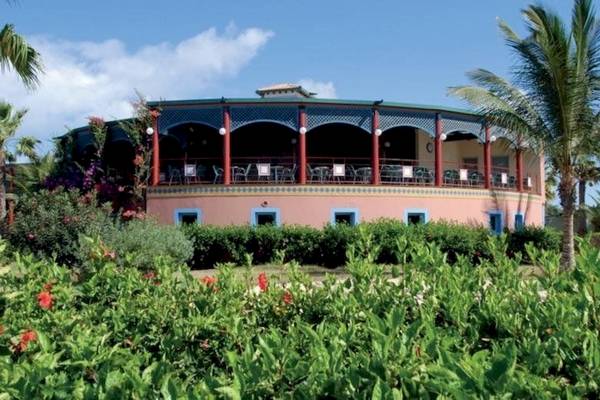 Außenansicht des VOI Vila Do Farol Resorts mit Terrasse, Tischen und Stühlen, umgeben von Pflanzen und Palmen.