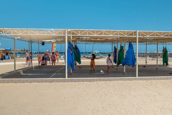 Ein Sonnensegel auf dem Strand mit mehreren Menschen, darunter Frauen in orangen Shorts, die vor einem Pavillon stehen.