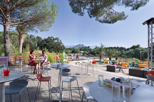 Terrasse des Belambra Club Golfe de Lozari Hotels mit Tischen und Stühlen unter Bäumen.