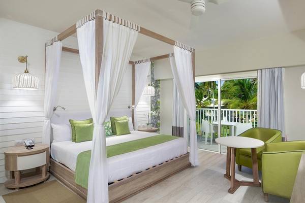 Innenansicht eines eleganten Zimmers im Grand Sirenis Punta Cana Resort mit Himmelbett, grünen Sesseln und Balkon.