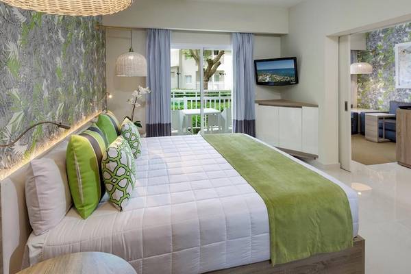 Innenansicht eines Schlafzimmers im Grand Sirenis Punta Cana Resort mit einem großen Bett, grünen Kissen und einem Fernseher an der Wand.