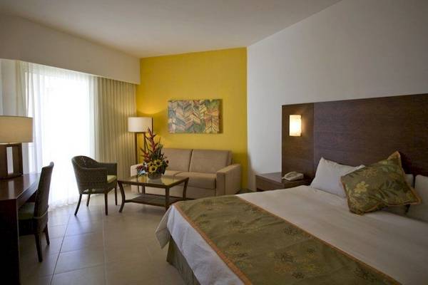Hotelzimmer im Dreams Royal Beach Punta Cana mit gelber Wand, Bett, Sofa, Tisch, Lampe und Fenster mit Vorhängen.