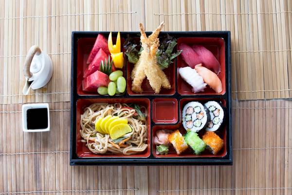 Asiatisches Essen in Schalen auf einer Bambusmatte mit verschiedenen Gerichten wie Sushi, Nudeln und Früchten.