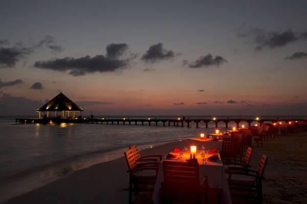 Ein abendlicher Blick auf das Coco Bodu Hithi Hotel, wo gesellige Tischplätze am Strand für ein romantisches Abendessen vorbereitet sind.