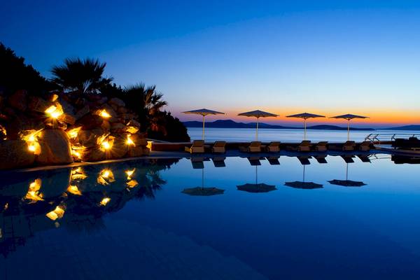 Pool im Mykonos Grand Hotel & Resort bei Sonnenuntergang mit beleuchteten Felsen und Sonnenschirmen.