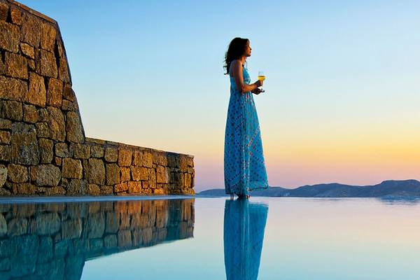 Frau im blauen Kleid am Pool am Abend vor einem steinernen Zaun und einer Aussicht auf Berge
