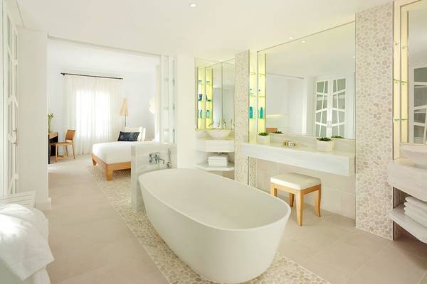 Badezimmer im Mykonos Grand Hotel & Resort mit Badewanne, Spiegel und offenem Bett im Hintergrund.