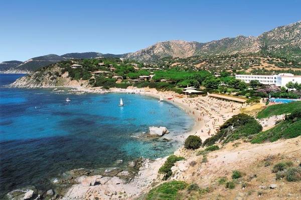 Luftaufnahme des Falkensteiner Resort Capo Boi Strandes mit vielen Besuchern, blauem Wasser und Bergen im Hintergrund.