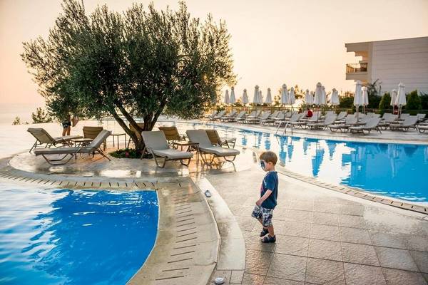 Ein Kind steht am Pool im Ikos Oceania Hotel neben einem Baum und vielen Liegen.