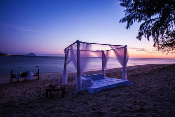 Ein Bett mit Baldachin auf dem Strand bei Sonnenuntergang mit einem Tisch davor.