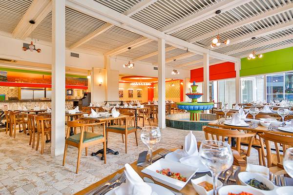 Innenraum des Restaurants im Crystal Admiral Resort Suites & Spa mit runden Tischen und Stühlen, einer zentralen Brunnen und bunten Wänden.