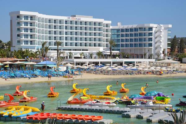Vassos Nissi Plage Hotel am Strand mit vielen bunten Wasserfahrzeugen und Sonnenschirmen.
