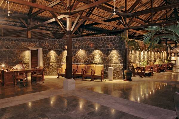 Innenansicht der Rezeption des Jimbaran Puri Hotels, Bali mit Steinwänden, Holzmöbeln und einem Schreibtisch.