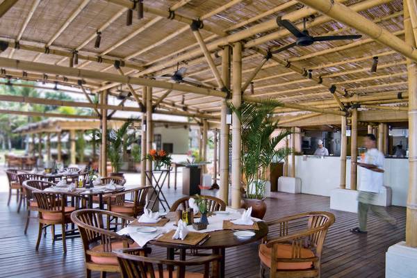 Innenbereich des Restaurants des Jimbaran Puri Hotels in Bali mit Holztischen und Stühlen unter einem Bambusdach.