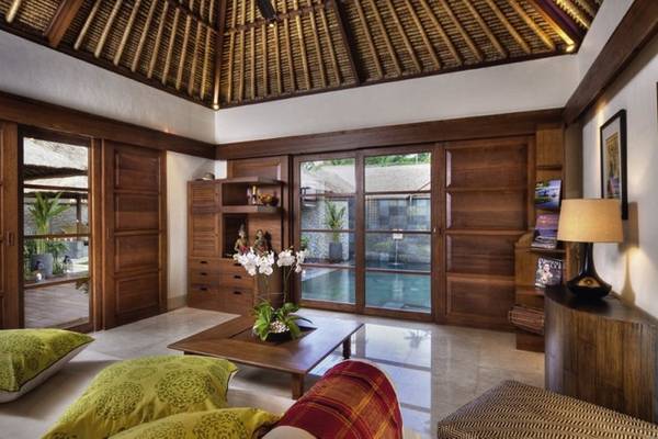 Wohnzimmer des Jimbaran Puri Hotels in Bali mit Holzmöbeln, einem Pool im Hintergrund und einer Blumenvase auf dem Tisch.
