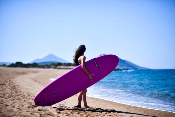 Eine Frau steht am Strand des Westin Resort Costa Navarino und hält ein pinkes Surfbrett.