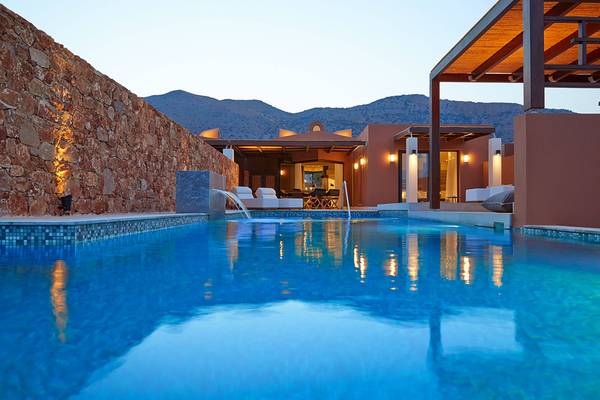 Der Pool des Domes of Elounda Hotels bei Dämmerung mit steinerner Wand und Bergen im Hintergrund.