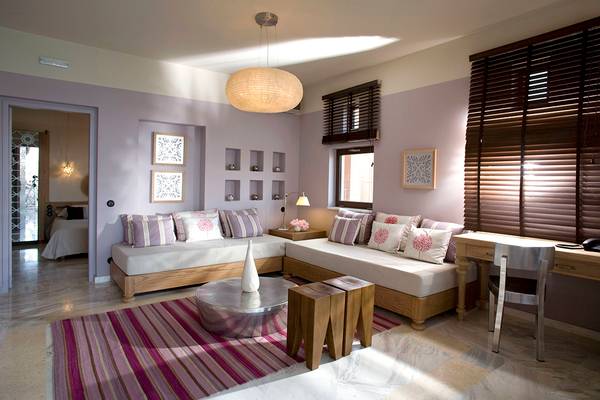 Suite-Schlafzimmer im Domes of Elounda mit zwei Sofas, Tisch, Stuhl, Lampe und Wanddekoration.