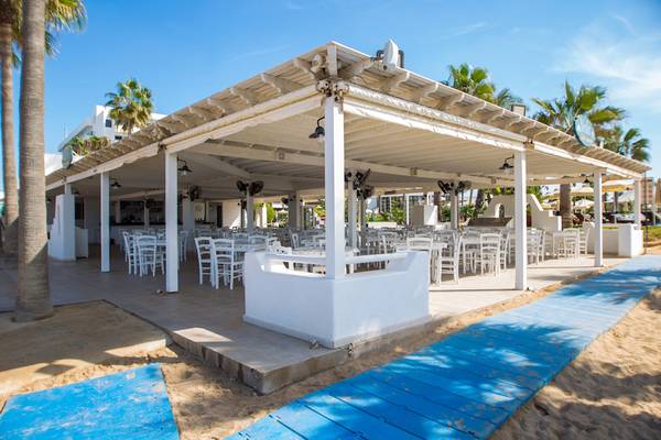 Terrasse des Adams Beach Hotels mit weißen Tischen und Stühlen unter einem Pergoladach am Strand.