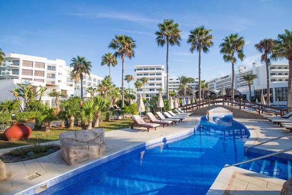 Außenpoolbereich des Adams Beach Hotels mit Palmen, Sonnenliegen und weißen Gebäuden im Hintergrund.