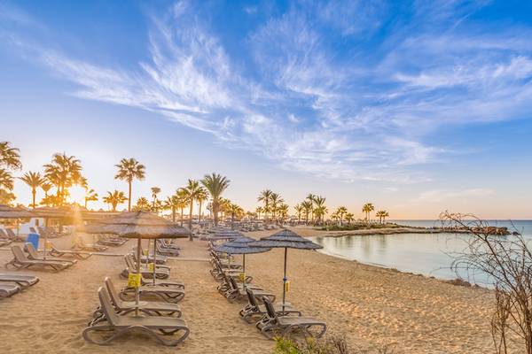 Strandansicht des Adams Beach Hotels mit Sonnenuntergang und Liegestühlen.