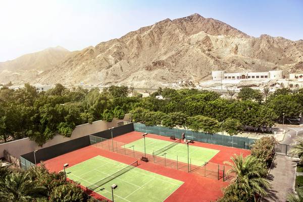 Tennisplatz im Fujairah Rotana Resort & Spa mit grünen Plätzen und roten Böden, umgeben von Bäumen und Bergen.