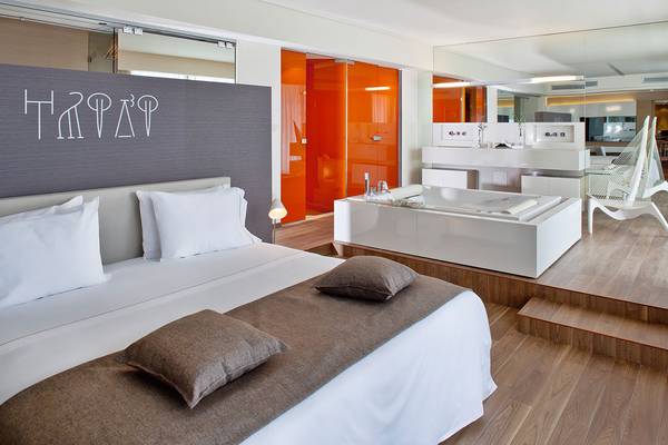 Schlafzimmer im Avra Imperial Hotel mit Bett, Kissen, Badewanne und Spiegel.